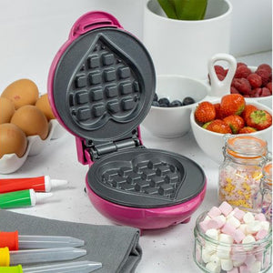 Pink heart-shaped waffle maker with non-stick plates open, surrounded by ingredients including eggs, berries, strawberries, blueberries, marshmallows in glass jars, a potted plant, and colorful markers on a white surface