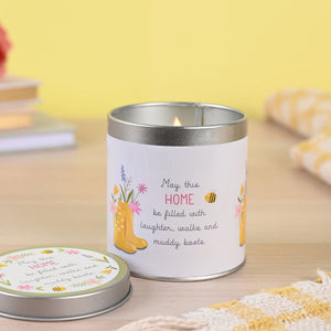 White tin candle with silver rim featuring yellow wellington boots, pink and blue flowers, and bee illustrations with text reading "May this HOME be filled with laughter, walks and muddy boots" displayed on wooden surface with matching lid nearby against yellow background
