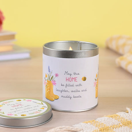White tin candle with silver rim featuring yellow wellington boots, pink and blue flowers, and bee illustrations with text reading "May this HOME be filled with laughter, walks and muddy boots" displayed on wooden surface with matching lid nearby against yellow background