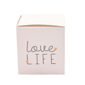Pale pink cylindrical tin candle with gray and coral text reading "love LIFE" decorated with small floral accents, displayed against a white background