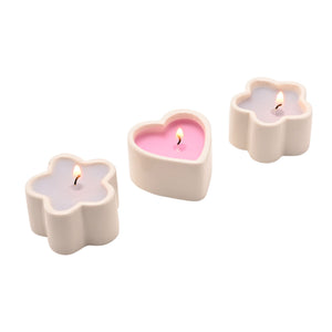 Three decorative mini scented candles in cream ceramic flower-shaped containers with pink and lavender wax, each with a lit flame, arranged in a diagonal line on a white background