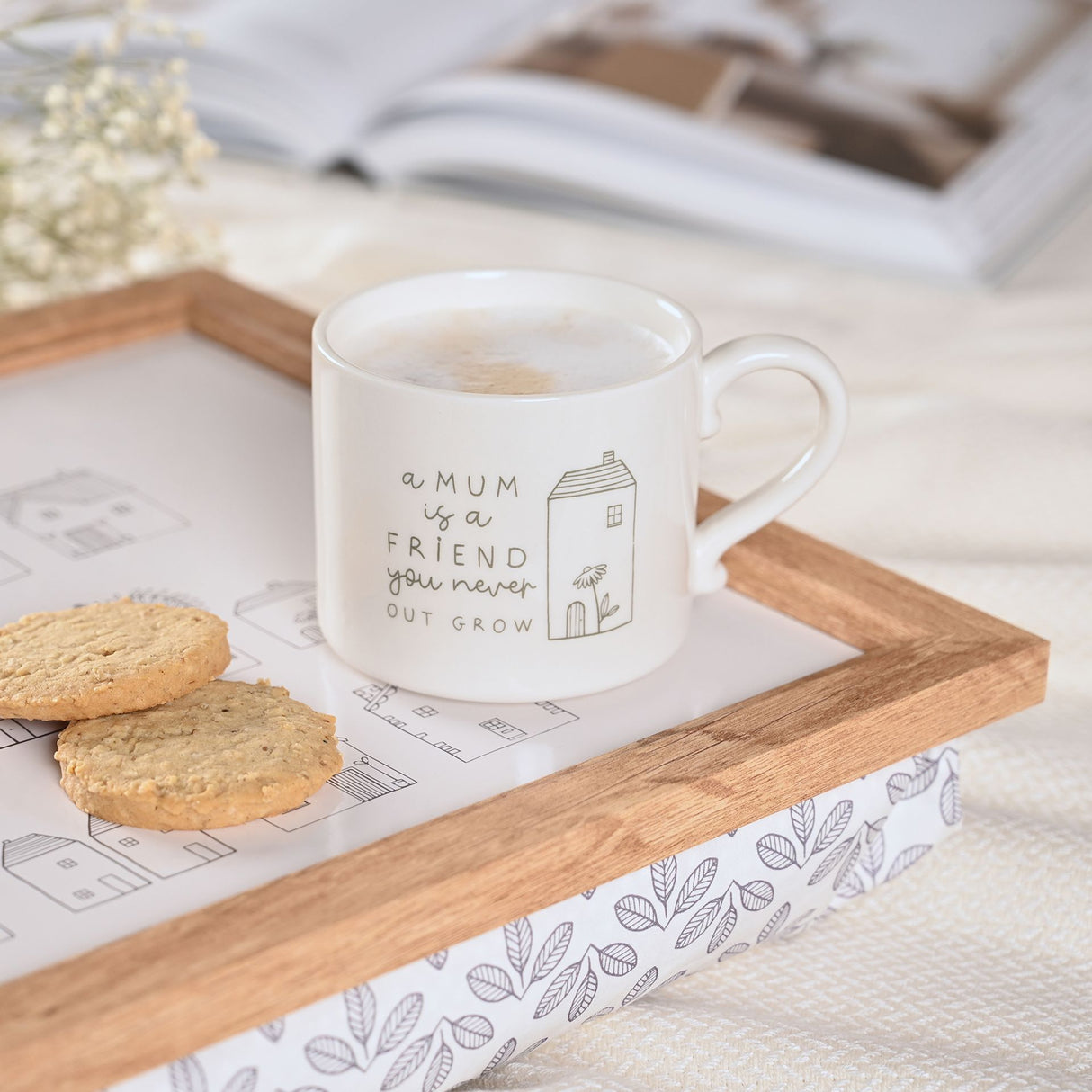 White ceramic mug with grey line illustration featuring text "a MUM is a FRIEND you never OUT GROW" and a small house design, filled with hot beverage, placed on wooden tray with oatmeal cookies and decorative leaf-patterned napkin