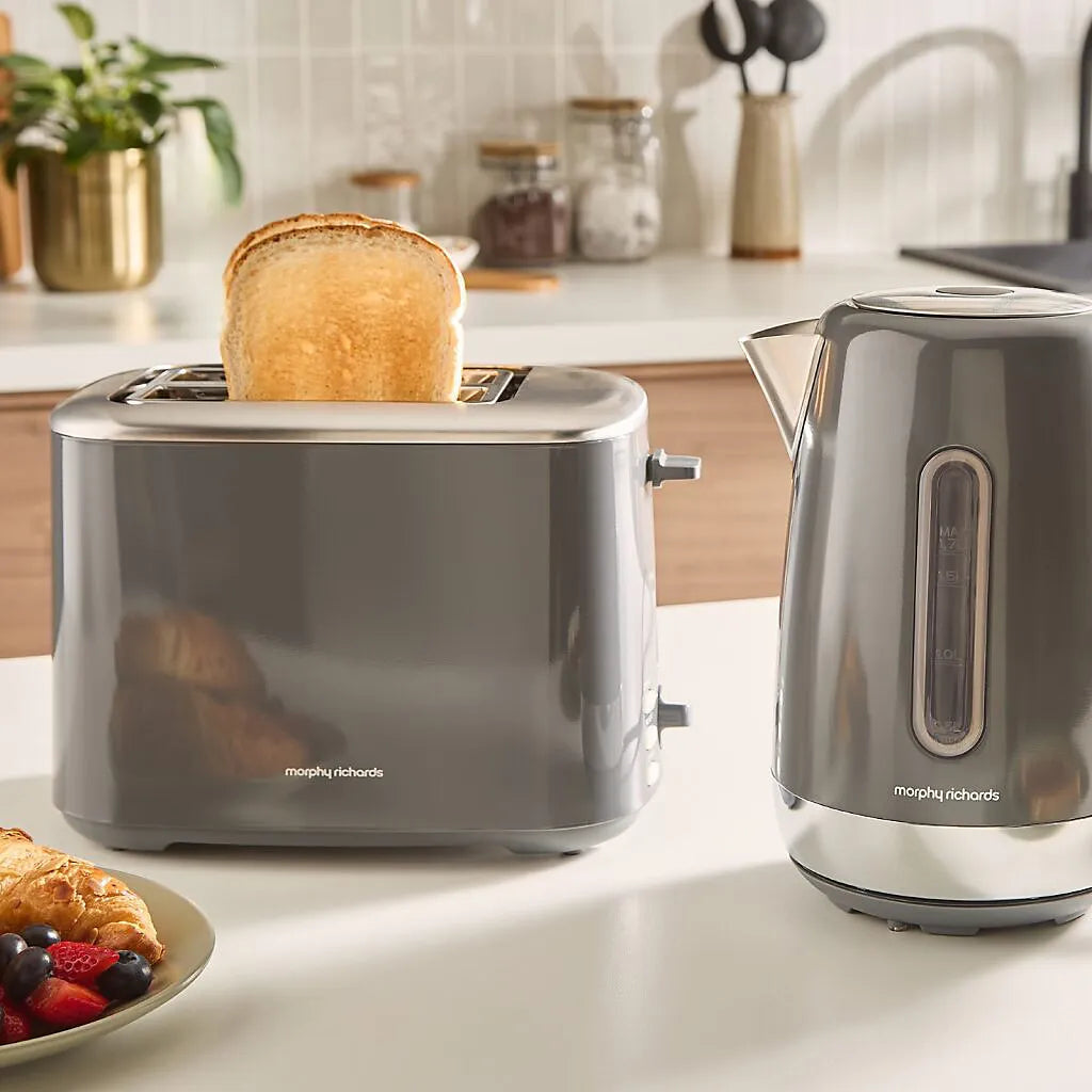 Morphy Richards Equip 2 slice toaster in grey stainless steel with a slice of golden toast popping up, displayed on a white kitchen countertop next to a matching grey electric kettle, with breakfast items and kitchen accessories in the background