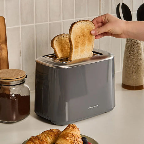 Morphy Richards Equip 2 slice toaster in grey with two slices of golden brown toast being removed, displayed on a white countertop with kitchen accessories including a jar of spread and wooden utensils in the background