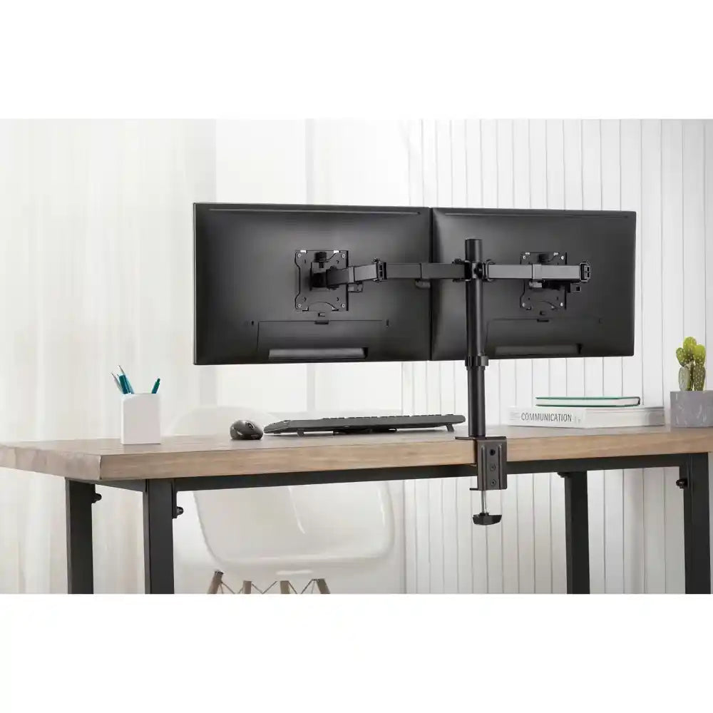 Black dual monitor clamp mount with articulating arms holding two black monitors above a wooden desk with black metal frame, alongside a white pen holder, keyboard, mouse and small potted plant in a bright office setting