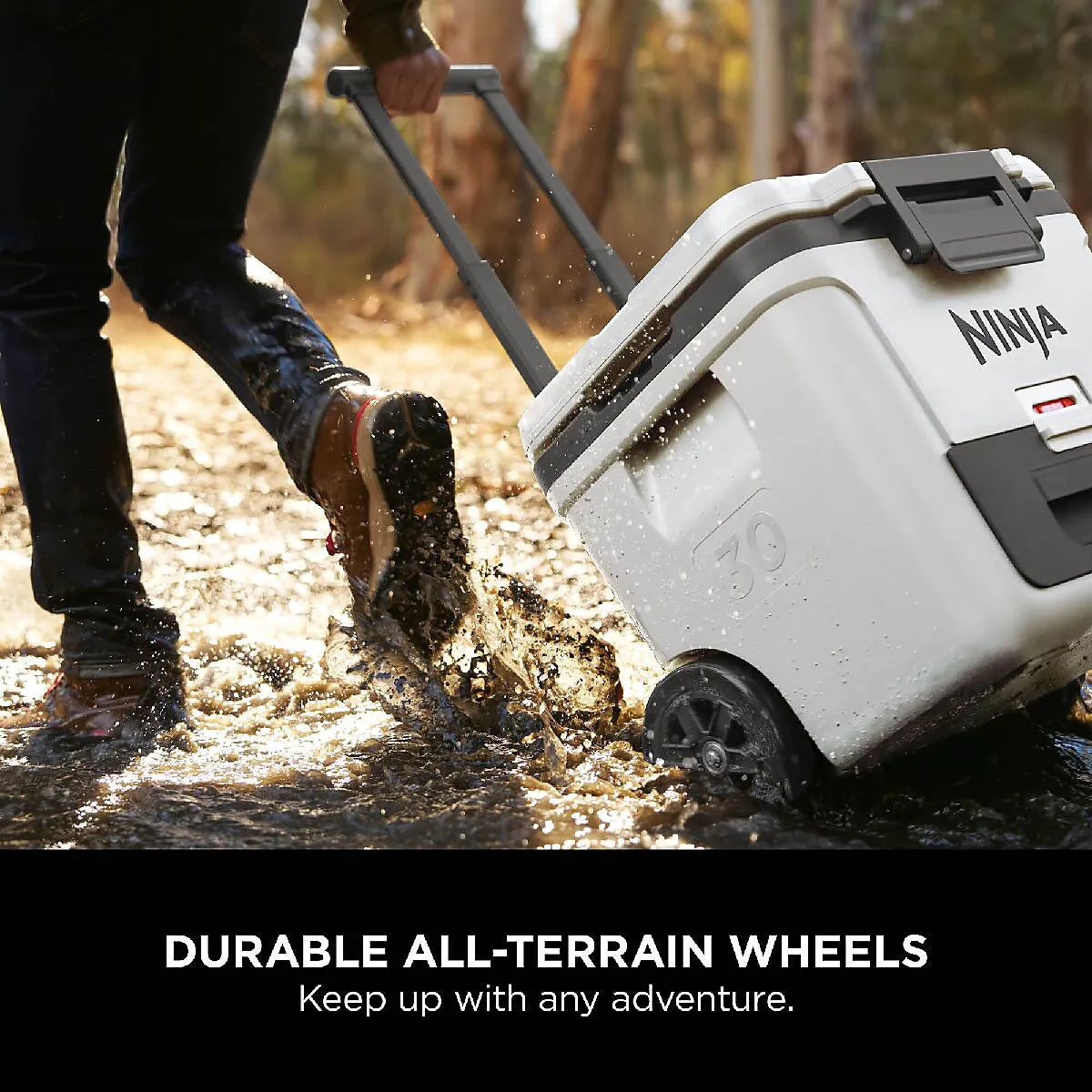 White Ninja FrostVault 28 litre wheeled cooler box with black handles being pulled through muddy water on a forest path, demonstrating durable all-terrain wheels for outdoor adventures