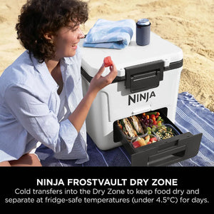 Woman in light blue shirt holding watermelon beside white Ninja FrostVault 28 litre wheeled cooler with black handles and open drawer showing fresh food items on sandy beach with towel and drink holder visible on top