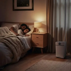 Masterplug Puredry SmartEnergy white dehumidifier with digital display on wooden bedroom floor next to bed with sleeping couple, warm lamp lighting and curtains visible