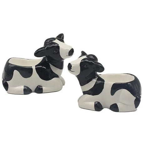 Set of two ceramic cow-shaped egg cups in black and white with glossy finish, featuring detailed facial features and lying down posture, displayed against white background
