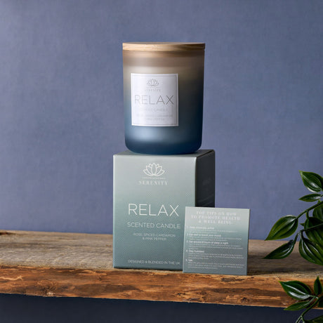 Serenity Relax scented candle in dark glass jar with wooden lid, displayed on rustic wooden surface with packaging box and wellness tips card, featuring rose cardamom and pink pepper fragrance, against blue-grey background with green foliage