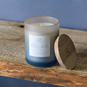 Serenity Relax scented candle in frosted glass jar with white label featuring lotus logo, wooden lid with engraved branding, displayed on rustic wooden surface against blue background, rose cardamom and pink pepper fragrance 120g