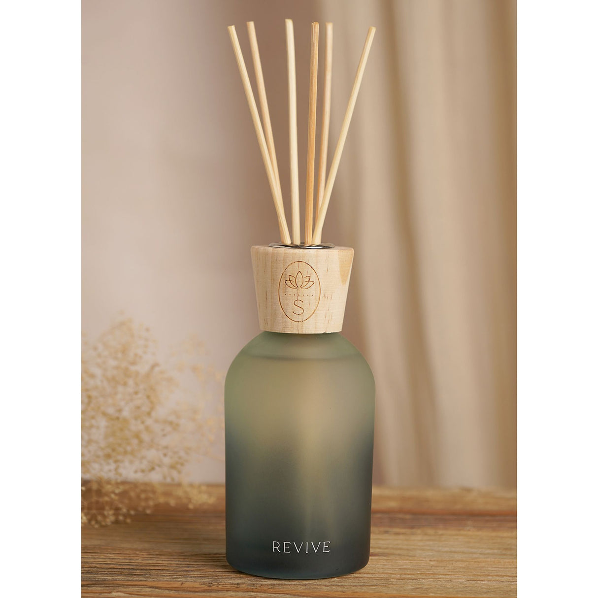 Reed diffuser with frosted sage green glass bottle, natural wooden cap with lotus engraved logo, six wooden diffuser sticks, labeled REVIVE, displayed on wooden surface against neutral beige curtain background