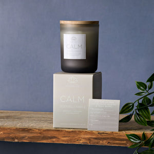 Black frosted glass scented candle with wooden lid labeled CALM by Serenity, displayed on cream packaging box with product information card, featuring bergamot lavender and sandalwood fragrance, arranged on rustic wooden surface against blue-gray background with green foliage