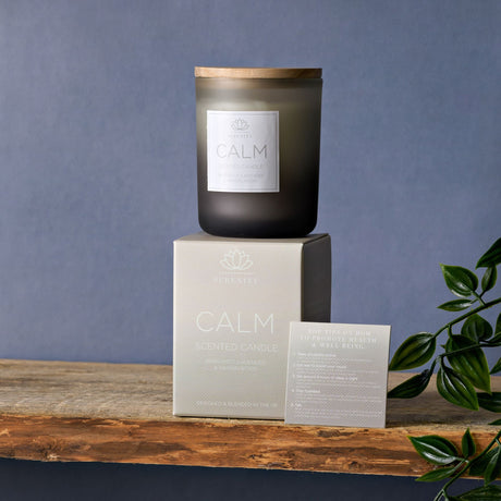 Black frosted glass scented candle with wooden lid labeled CALM by Serenity, displayed on cream packaging box with product information card, featuring bergamot lavender and sandalwood fragrance, arranged on rustic wooden surface against blue-gray background with green foliage