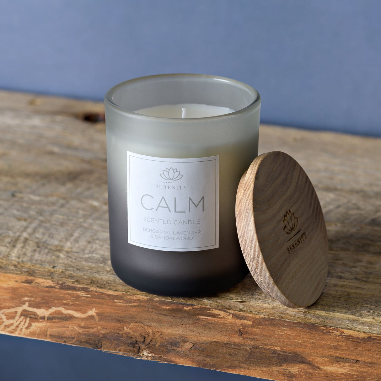 Serenity Calm scented candle in frosted glass jar with white label featuring lotus logo, accompanied by wooden lid with engraved branding, displayed on rustic driftwood against blue background