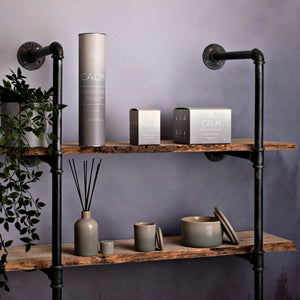 Industrial-style black metal pipe shelving unit with reclaimed wooden shelves displaying the Serenity Calm candle collection in cream and charcoal packaging, including bergamot lavender and sandalwood scented candles, diffusers, and ceramic vessels against a purple-grey wall with green foliage