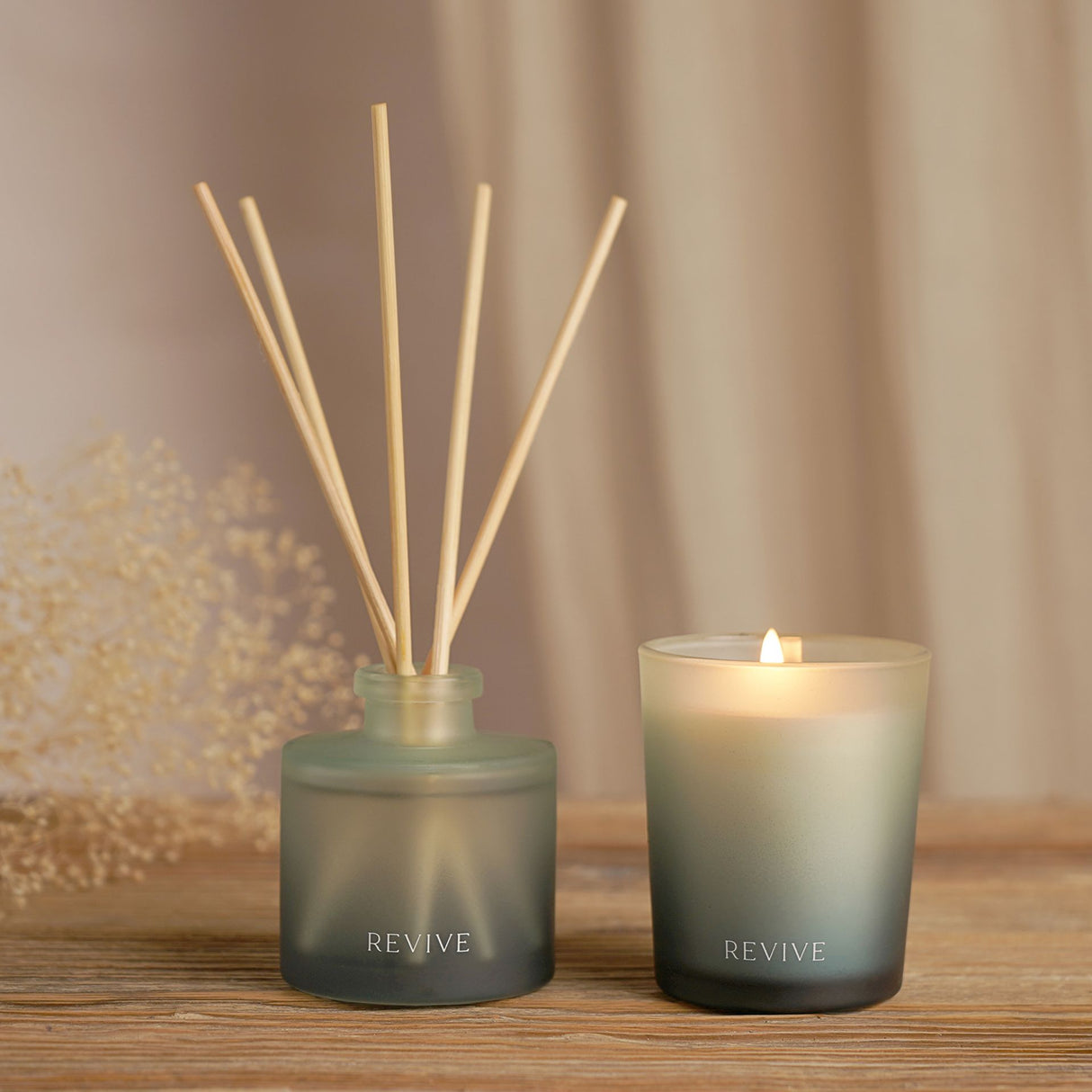 Serenity Revive luxury candle and diffuser set featuring a frosted sage green glass candle with lit flame and matching reed diffuser with natural wooden sticks on wooden surface against neutral beige curtain backdrop