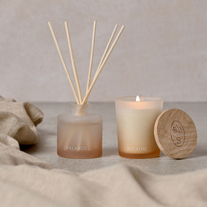 Serenity Breathe Diffuser and Candle Set featuring a reed diffuser labeled BALANCE with natural wooden sticks, a cream candle labeled BREATHE with lit flame, and wooden lid with lotus engraving, arranged on neutral linen fabric against a soft beige background