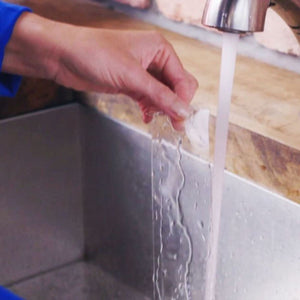 Hand demonstrating double-sided adhesive tape application on white tape roll, showing waterproof bonding on kitchen sink edge with faucet visible in background