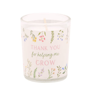A frosted glass candle with delicate watercolor floral illustrations in pink, yellow, purple and green featuring hanging flowers and botanical elements, with pink and sage green text reading "THANK YOU for helping me GROW" on a cream background