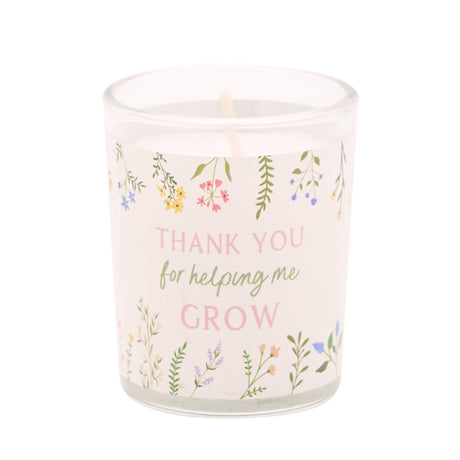 A frosted glass candle with delicate watercolor floral illustrations in pink, yellow, purple and green featuring hanging flowers and botanical elements, with pink and sage green text reading "THANK YOU for helping me GROW" on a cream background