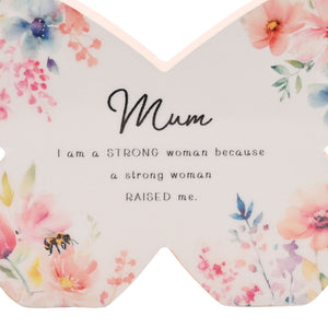 Decorative butterfly-shaped ceramic mantel plaque with watercolor floral design featuring pink, blue and purple flowers, green leaves and a bee, inscribed with "Mum" in cursive script and the message "I am a STRONG woman because a strong woman RAISED me" in black text on a cream background