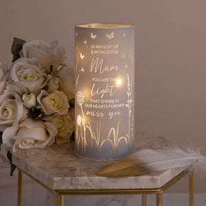 Frosted glass tube light with gold engraved text reading "In Memory of a Wonderful Mum, You Are The Light That Shines in Our Hearts Forever, Miss You" decorated with butterflies and dandelions, illuminated from within, displayed on a marble hexagonal table next to a bouquet of cream and white roses