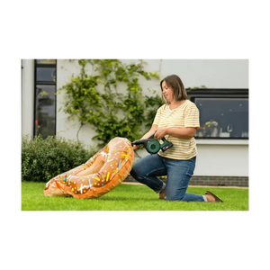Woman using a Bosch cordless leaf blower on grass in a garden, wearing a yellow and white striped t-shirt and blue jeans, with an inflatable orange dragon toy nearby and a modern house with ivy in the background