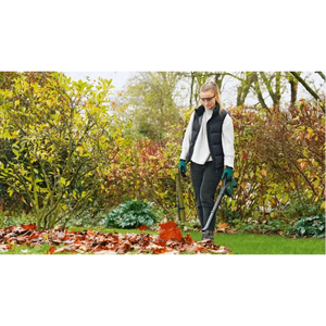 Woman using Bosch Universal Garden Leaf Blower cordless 18V to clear fallen autumn leaves from lawn surrounded by hedges and trees in garden setting