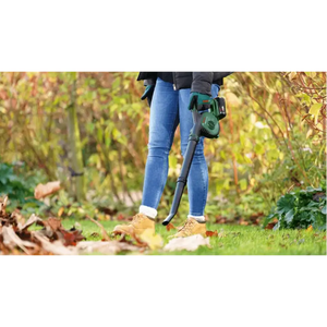 Person wearing blue jeans and tan boots using a Bosch cordless leaf blower with green and black design in an autumn garden surrounded by fallen leaves and shrubs
