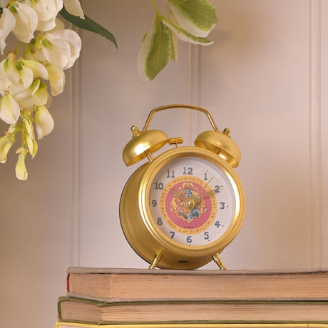 Gold twin bell alarm clock with Hogwarts crest on white dial, sitting on stacked books with white and yellow flowers above against pale pink wall
