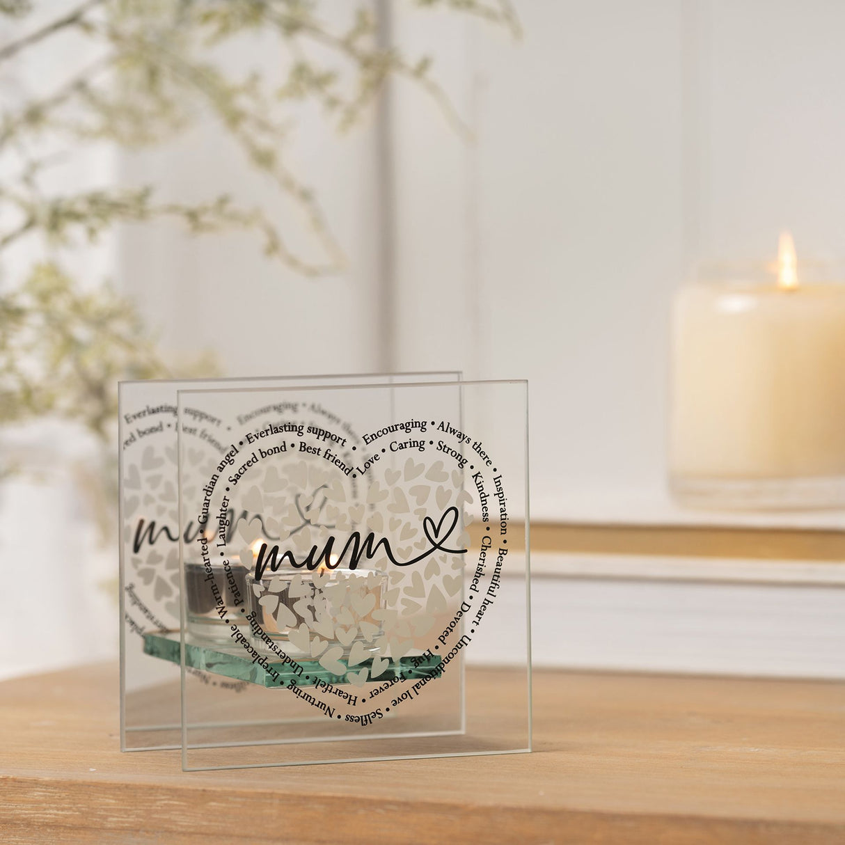 Decorative glass item with 'mum' and heart design on a wooden surface with a blurred candle and plant in the background.