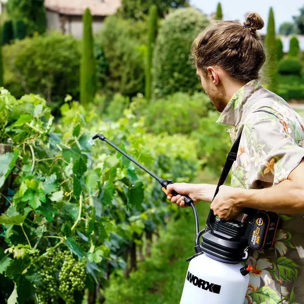 Man using WorX 20V cordless sprayer in lush green garden with vegetables and cypress hedges, demonstrating pesticide or fertilizer application on plant foliage
